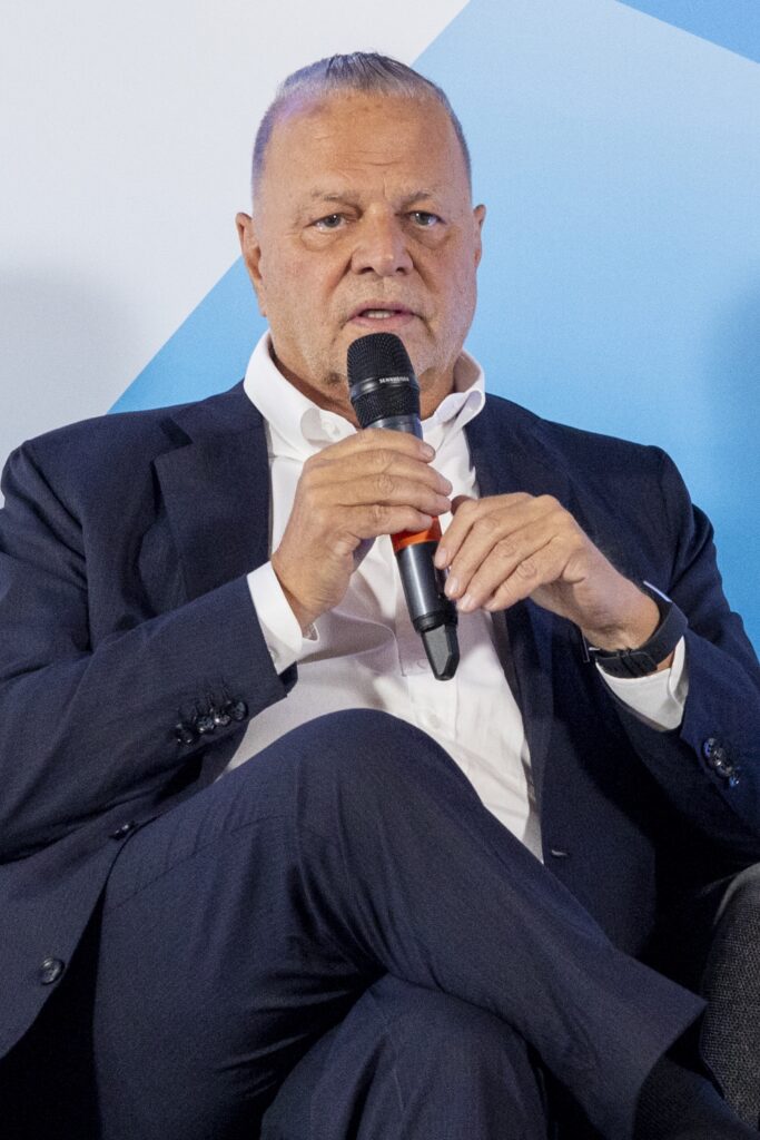 A man in a suit holds a microphone while speaking, seated against a blue background.