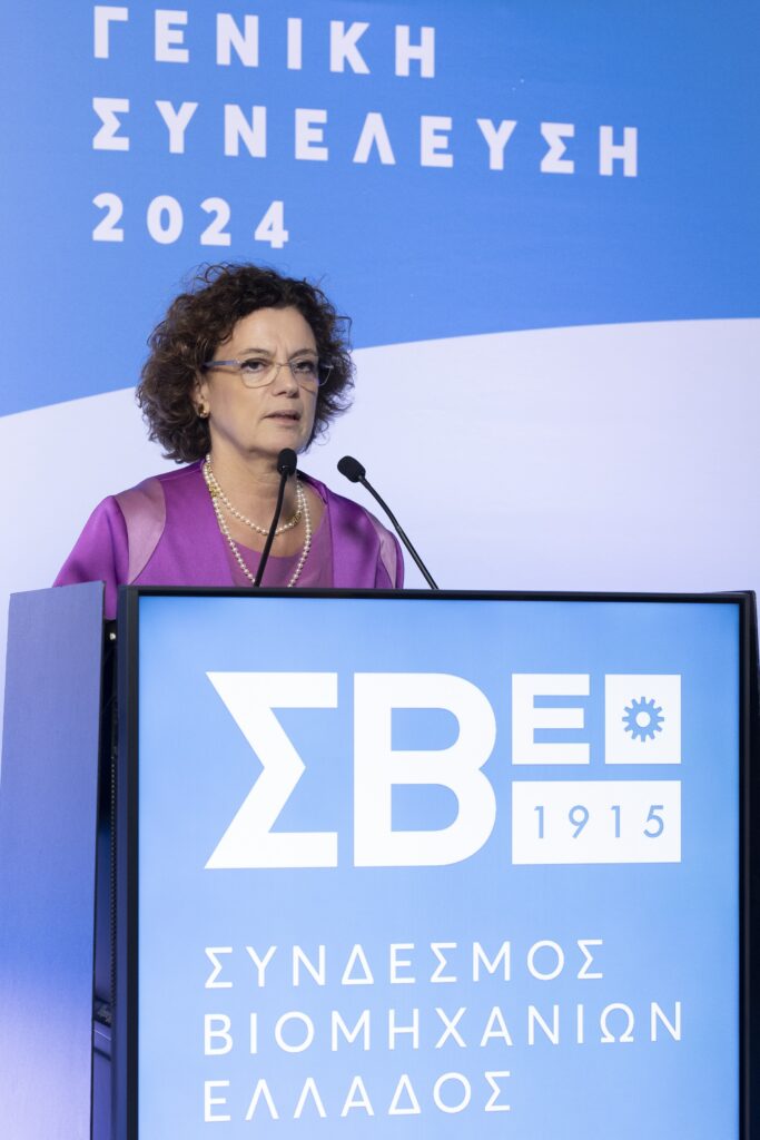 A woman in a purple outfit speaks at a podium with a blue backdrop, featuring Greek text and a logo.