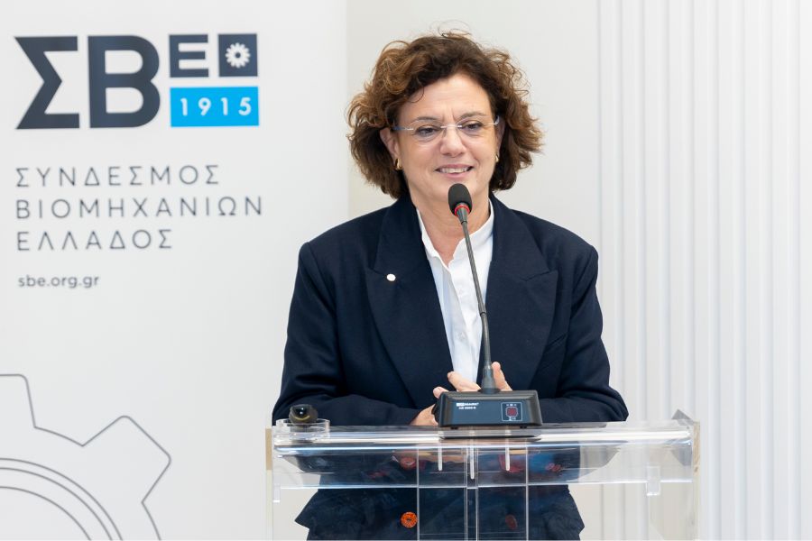 A woman with curly hair speaks at a podium, wearing a dark blazer, in front of a backdrop featuring a logo and Greek text.