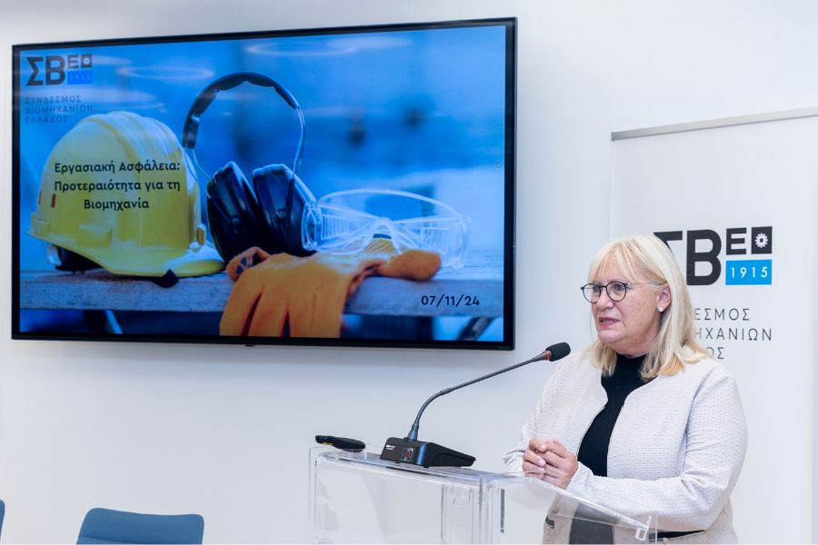 A woman speaks at a podium with a presentation screen behind her, displaying safety equipment for industry.